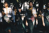 A group of people wearing uniforms adorned with numerous badges, indicating some form of achievement or participation, possibly related to scouting or similar organizations. The scene is set outdoors in a sunlit park area with trees providing shade. The individuals appear to be walking in a relaxed and friendly manner.
