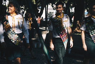 A group of people wearing uniforms adorned with numerous badges, indicating some form of achievement or participation, possibly related to scouting or similar organizations. The scene is set outdoors in a sunlit park area with trees providing shade. The individuals appear to be walking in a relaxed and friendly manner.