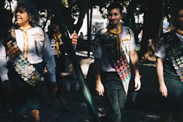 A group of people wearing uniforms adorned with numerous badges, indicating some form of achievement or participation, possibly related to scouting or similar organizations. The scene is set outdoors in a sunlit park area with trees providing shade. The individuals appear to be walking in a relaxed and friendly manner.