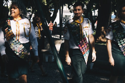 A group of people wearing uniforms adorned with numerous badges, indicating some form of achievement or participation, possibly related to scouting or similar organizations. The scene is set outdoors in a sunlit park area with trees providing shade. The individuals appear to be walking in a relaxed and friendly manner.