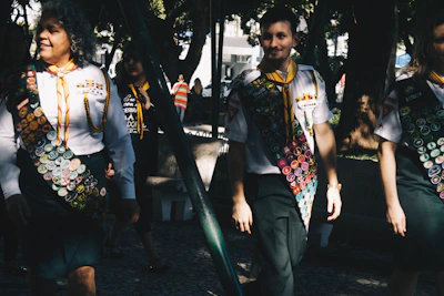 A group of people wearing uniforms adorned with numerous badges, indicating some form of achievement or participation, possibly related to scouting or similar organizations. The scene is set outdoors in a sunlit park area with trees providing shade. The individuals appear to be walking in a relaxed and friendly manner.