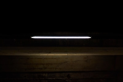 A long, illuminated fluorescent light tube is mounted against a dark background, casting a faint glow onto a rough, textured concrete surface below.