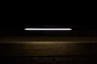 A long, illuminated fluorescent light tube is mounted against a dark background, casting a faint glow onto a rough, textured concrete surface below.