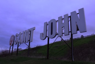Large white letters spelling 'SAINT JOHN' are mounted on a hillside. The background features a cloudy sky with a purple tint. The letters are supported by a metal frame, and there is a grassy slope beneath.