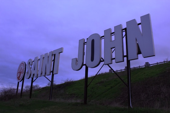 Large white letters spelling 'SAINT JOHN' are mounted on a hillside. The background features a cloudy sky with a purple tint. The letters are supported by a metal frame, and there is a grassy slope beneath.