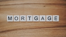 The word 'MORTGAGE' is spelled out using white letter tiles, each with black uppercase letters, arranged in a row on a wooden surface.