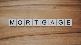 The word 'MORTGAGE' is spelled out using white letter tiles, each with black uppercase letters, arranged in a row on a wooden surface.