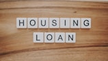 Wooden background features a set of letter tiles spelling out the phrase 'HOUSING LOAN' in two rows. The tiles have black letters on a white surface, creating a simple and clear message.