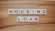 Wooden background features a set of letter tiles spelling out the phrase 'HOUSING LOAN' in two rows. The tiles have black letters on a white surface, creating a simple and clear message.