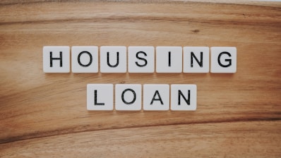 Wooden background features a set of letter tiles spelling out the phrase 'HOUSING LOAN' in two rows. The tiles have black letters on a white surface, creating a simple and clear message.
