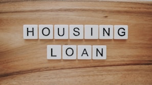 Wooden background features a set of letter tiles spelling out the phrase 'HOUSING LOAN' in two rows. The tiles have black letters on a white surface, creating a simple and clear message.