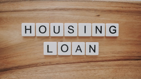 Wooden background features a set of letter tiles spelling out the phrase 'HOUSING LOAN' in two rows. The tiles have black letters on a white surface, creating a simple and clear message.