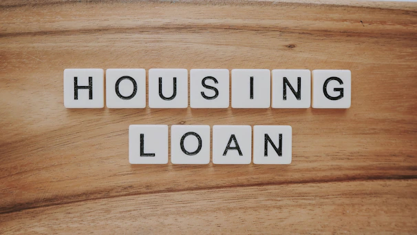 Wooden background features a set of letter tiles spelling out the phrase 'HOUSING LOAN' in two rows. The tiles have black letters on a white surface, creating a simple and clear message.