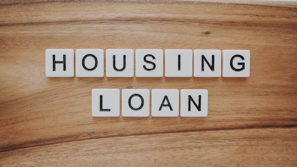 Wooden background features a set of letter tiles spelling out the phrase 'HOUSING LOAN' in two rows. The tiles have black letters on a white surface, creating a simple and clear message.