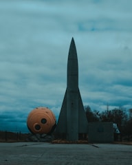 A large, gray rocket stands upright on a concrete surface with a spherical orange object lying beside it. The sky is overcast with various shades of blue and gray clouds. Trees can be seen in the background.