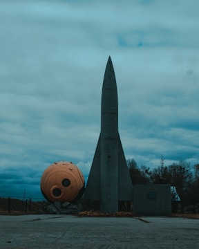 A large, gray rocket stands upright on a concrete surface with a spherical orange object lying beside it. The sky is overcast with various shades of blue and gray clouds. Trees can be seen in the background.