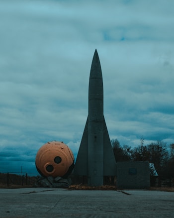 A large, gray rocket stands upright on a concrete surface with a spherical orange object lying beside it. The sky is overcast with various shades of blue and gray clouds. Trees can be seen in the background.
