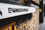 A graffiti-style stencil with the text '@iShootHumans' is painted on a white surface attached to a yellow brick wall. The focus is on the stencil while the background, consisting of more of the brick wall and a blurry street view, is out of focus.