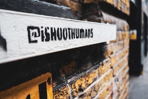 A graffiti-style stencil with the text '@iShootHumans' is painted on a white surface attached to a yellow brick wall. The focus is on the stencil while the background, consisting of more of the brick wall and a blurry street view, is out of focus.