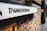 A graffiti-style stencil with the text '@iShootHumans' is painted on a white surface attached to a yellow brick wall. The focus is on the stencil while the background, consisting of more of the brick wall and a blurry street view, is out of focus.