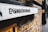 A graffiti-style stencil with the text '@iShootHumans' is painted on a white surface attached to a yellow brick wall. The focus is on the stencil while the background, consisting of more of the brick wall and a blurry street view, is out of focus.