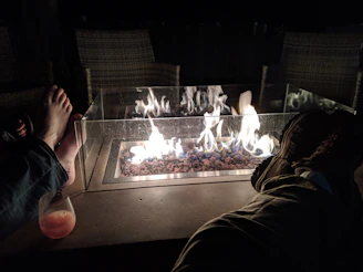 A fire pit with flames burning over stones is set in the center. Two pairs of feet are visible, one bare and the other wearing shoes, suggesting people lounging around the fire. A glass panel surrounds the fire, and a drink is placed on the surface beside one pair of feet. The setting appears cozy and relaxed, likely outdoors at night with chairs in the background.
