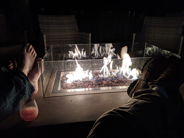 A fire pit with flames burning over stones is set in the center. Two pairs of feet are visible, one bare and the other wearing shoes, suggesting people lounging around the fire. A glass panel surrounds the fire, and a drink is placed on the surface beside one pair of feet. The setting appears cozy and relaxed, likely outdoors at night with chairs in the background.