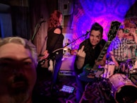 A live music performance with three individuals on stage. One person with a guitar is making a gesture towards the camera, while another stands by a microphone. The third person is seated at a drum set. The lighting is colorful and dynamic, creating a lively atmosphere.