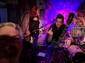 A live music performance with three individuals on stage. One person with a guitar is making a gesture towards the camera, while another stands by a microphone. The third person is seated at a drum set. The lighting is colorful and dynamic, creating a lively atmosphere.