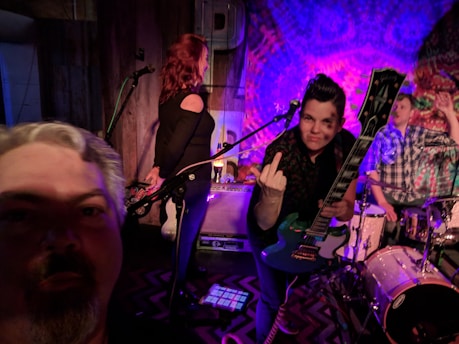 A live music performance with three individuals on stage. One person with a guitar is making a gesture towards the camera, while another stands by a microphone. The third person is seated at a drum set. The lighting is colorful and dynamic, creating a lively atmosphere.