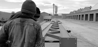 Several individuals in work attire are observing steel beams in an industrial setting, with storage buildings on one side and a silo in the background.