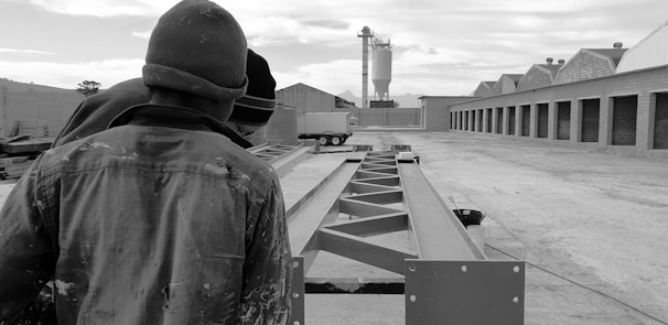 Several individuals in work attire are observing steel beams in an industrial setting, with storage buildings on one side and a silo in the background.