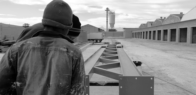 Several individuals in work attire are observing steel beams in an industrial setting, with storage buildings on one side and a silo in the background.