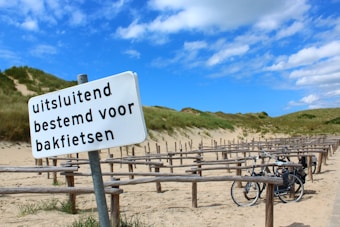 A sandy path with a row of wooden bike racks is visible, surrounded by grassy dunes under a partly cloudy blue sky. A sign in Dutch, 'uitsluitend bestemd voor bakfietsen,' indicates it is designated for cargo bikes. Two bikes are parked at the racks.