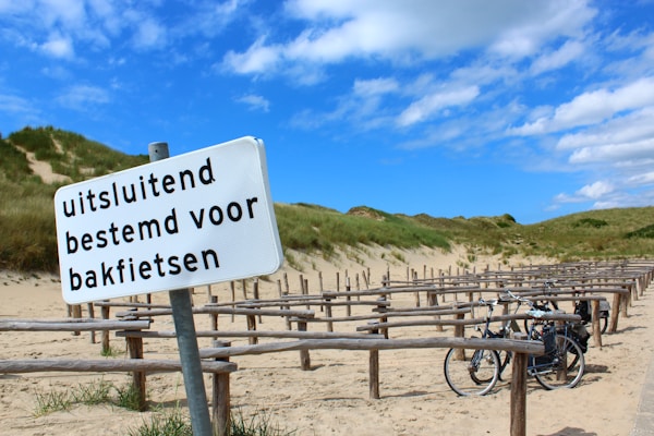 A sandy path with a row of wooden bike racks is visible, surrounded by grassy dunes under a partly cloudy blue sky. A sign in Dutch, 'uitsluitend bestemd voor bakfietsen,' indicates it is designated for cargo bikes. Two bikes are parked at the racks.