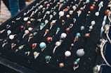 A display of numerous handcrafted rings, each featuring a unique, colorful gemstone set in silver wire wrapping, arranged neatly on a black velvet board. The stones vary in color and pattern, creating an artistic and vibrant presentation.