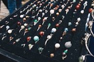 A display of numerous handcrafted rings, each featuring a unique, colorful gemstone set in silver wire wrapping, arranged neatly on a black velvet board. The stones vary in color and pattern, creating an artistic and vibrant presentation.