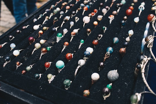 A display of numerous handcrafted rings, each featuring a unique, colorful gemstone set in silver wire wrapping, arranged neatly on a black velvet board. The stones vary in color and pattern, creating an artistic and vibrant presentation.