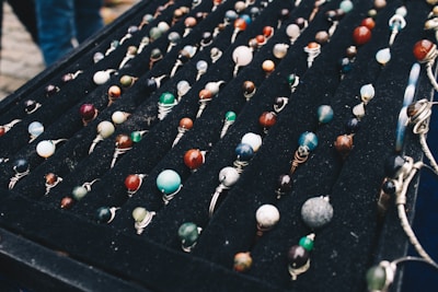 A display of numerous handcrafted rings, each featuring a unique, colorful gemstone set in silver wire wrapping, arranged neatly on a black velvet board. The stones vary in color and pattern, creating an artistic and vibrant presentation.