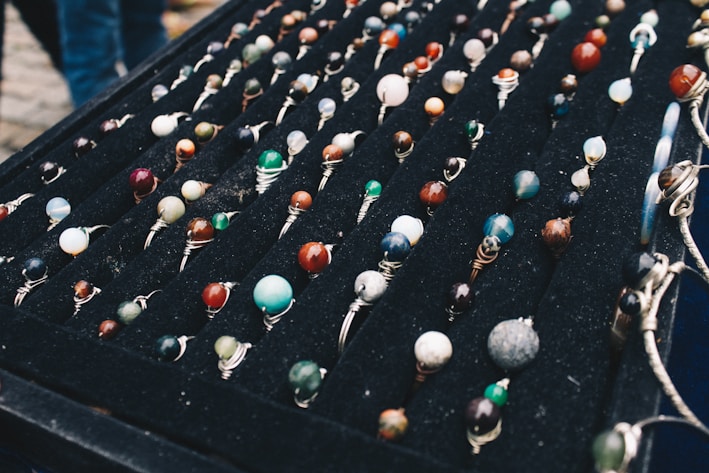 A display of numerous handcrafted rings, each featuring a unique, colorful gemstone set in silver wire wrapping, arranged neatly on a black velvet board. The stones vary in color and pattern, creating an artistic and vibrant presentation.