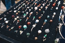 A display of numerous handcrafted rings, each featuring a unique, colorful gemstone set in silver wire wrapping, arranged neatly on a black velvet board. The stones vary in color and pattern, creating an artistic and vibrant presentation.