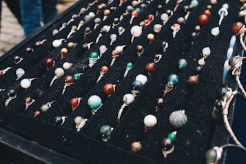 A display of numerous handcrafted rings, each featuring a unique, colorful gemstone set in silver wire wrapping, arranged neatly on a black velvet board. The stones vary in color and pattern, creating an artistic and vibrant presentation.