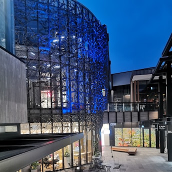 A modern architectural space with intricate metal lattice work illuminated by blue lighting on the facade of a building. The area is a commercial or office environment with glass windows revealing an interior space bustling with activity. There are outdoor seating arrangements and decorative plants scattered around a pedestrian pathway.