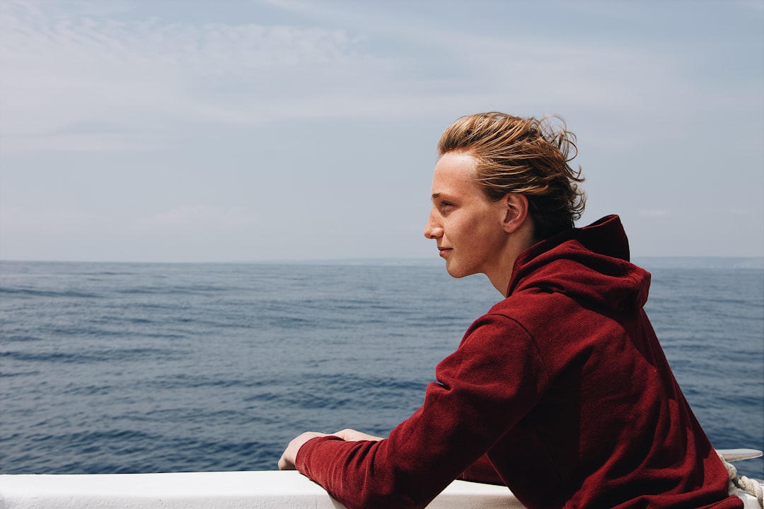 man in maroon hoodie leaning on wall overlooking sea