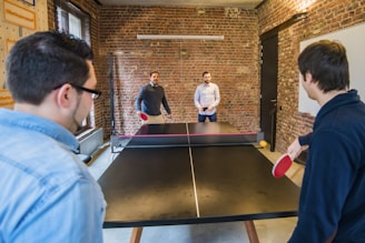 Four people engage in a ping pong game in a brick-walled room. Two players are actively playing while the other two observe from the other side. The room features a casual setting with a large window on one side and a whiteboard on the opposite wall.