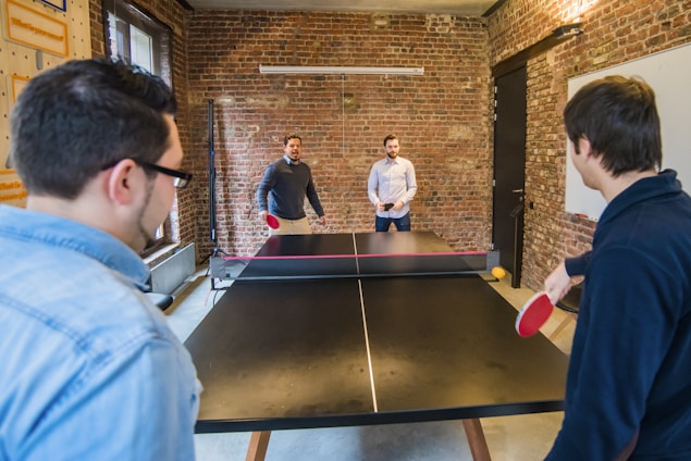 Four people engage in a ping pong game in a brick-walled room. Two players are actively playing while the other two observe from the other side. The room features a casual setting with a large window on one side and a whiteboard on the opposite wall.