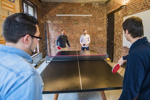 Four people engage in a ping pong game in a brick-walled room. Two players are actively playing while the other two observe from the other side. The room features a casual setting with a large window on one side and a whiteboard on the opposite wall.