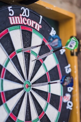 A close-up view of a dartboard with several darts embedded in it. The dartboard displays various colored sections, including green, red, white, and black, and features numerical values around the edge. The brand name 'unicorn' is visible at the top. The darts are striking the board at different angles.