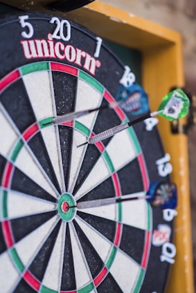 A close-up view of a dartboard with several darts embedded in it. The dartboard displays various colored sections, including green, red, white, and black, and features numerical values around the edge. The brand name 'unicorn' is visible at the top. The darts are striking the board at different angles.