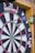A close-up view of a dartboard with several darts embedded in it. The dartboard displays various colored sections, including green, red, white, and black, and features numerical values around the edge. The brand name 'unicorn' is visible at the top. The darts are striking the board at different angles.